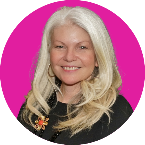 Portrait of a smiling woman with long silver hair, wearing a black top and a jeweled brooch, against a bright pink circular background.