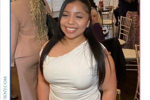 Smiling woman in a white one-shoulder top posing at a formal event with guests in the background and a 'Top Lawyers' frame above.