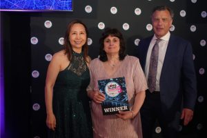 Three adults pose on a backdrop at an awards event; center woman holds a 'WINNER' plaque.