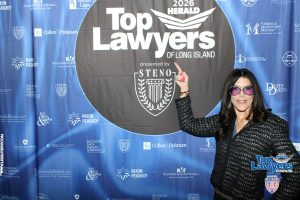 Smiling woman in a black jacket points to a large 'Top Lawyers of Long Island' banner at a law event backdrop.
