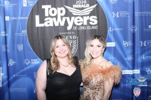 Two smiling women in formal dresses pose in front of a blue 'Top Lawyers of Long Island' backdrop at a 2026 event on the red carpet: one in black, one in leopard print.