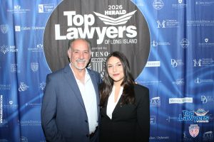 Two smiling adults pose in front of a blue sponsor-filled backdrop reading 'Top Lawyers of Long Island' at a 2026 event.