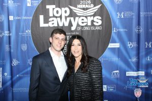 Man in a navy suit and woman in a black patterned jacket pose together in front of a blue 'Top Lawyers of Long Island' backdrop at a 2026 event.