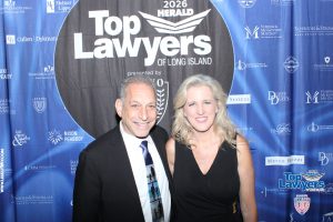 Smiling man in a tuxedo and a smiling woman in a black dress posing together on a blue step-and-repeat backdrop that reads 'Top Lawyers of Long Island' and is covered with sponsor logos.