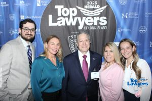 Five professionally dressed people pose together in front of a blue sponsor backdrop reading 'Top Lawyers of Long Island' at a formal event.