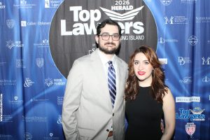 Two people posing in front of a 'Top Lawyers' backdrop at a formal event, the man in a light gray suit and the woman in a black dress smiling at the camera.