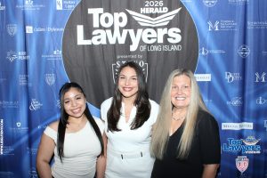 Three smiling women pose together in front of a blue banner that reads 'Top Lawyers of Long Island' at a 2026 Herald event.