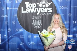 Woman in a pale pink dress holding a bouquet, posing in front of a 'Top Lawyers of Long Island' backdrop at a 2026 event.