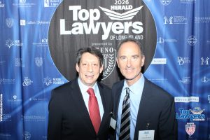 Two men in suits posing for a photo at a 2026 Top Lawyers event with a blue sponsor backdrop behind them