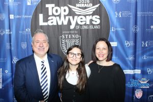 Three people pose and smile in front of a blue 'Top Lawyers of Long Island' backdrop featuring sponsor logos; a man in a suit left, a young woman with glasses center, and a woman in black on the right.