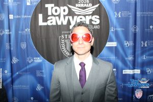 Man in a gray suit and purple tie wearing oversized pink heart-shaped sunglasses at a blue 'Top Lawyers' event backdrop.