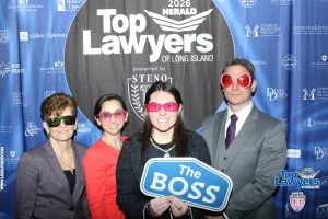 Group of four adults at a Top Lawyers event posing in front of a blue backdrop; central woman holds a 'The Boss' sign and wears pink sunglasses.