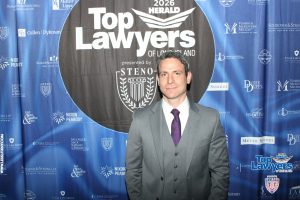Man in a gray suit and purple tie posing in front of a 'Top Lawyers' event backdrop with sponsor logos