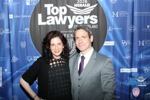 Two smiling adults posing in front of a blue 'Top Lawyers of Long Island' backdrop at a 2026 event, the woman in a black dress and the man in a gray suit.