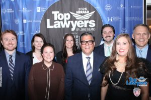 Group of nine professionally dressed people posing for a photo in front of a blue sponsor backdrop that reads 'Top Lawyers of Long Island' at the 2026 Herald event.