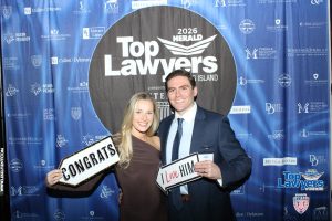Two smiling attendees pose in front of a blue 'Top Lawyers' backdrop, holding 'Congrats' and 'I Love Him' signs at a recognition event.