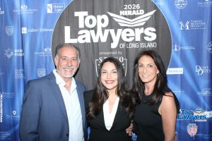 Three people smiling and posing in front of a blue "Top Lawyers of Long Island" backdrop at a 2026 event. In the center a woman in a black blazer stands with a man in a blue suit on her left and a woman in a black dress on her right.