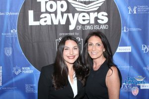 Two smiling women pose together at a 'Top Lawyers of Long Island' event in front of a blue sponsor backdrop.