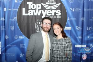 Two people posing in front of a 'Top Lawyers' event backdrop: man in a gray suit with a yellow tie and a woman in a black-and-white checkered dress.