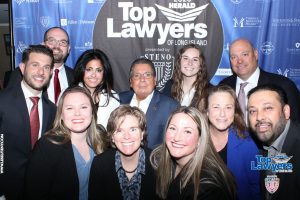Group of professionals in business attire posing for a group photo at a 'Top Lawyers of Long Island' event, with a blue sponsor banner behind them.