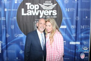 Smiling man in a suit and woman in a pink plaid shirt pose together in front of a blue 'Top Lawyers of Long Island' backdrop.
