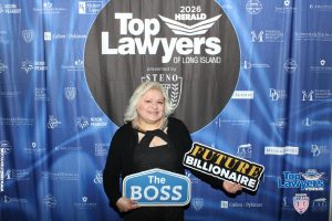 Smiling woman at Top Lawyers of Long Island event, holding a sign reading 'The Boss' with a 'Future Billionaire' cutout against a blue sponsor backdrop.