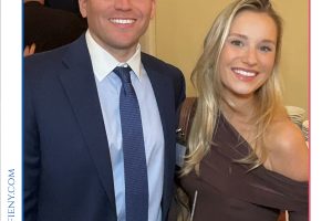 Smiling man in a navy suit and tie stands beside a blonde woman in a brown off‑the‑shoulder dress at a formal event, each holding a drink.