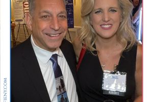 Two smiling adults in formal attire pose for a photo at an awards event, one man in a suit and tie and a woman in a black dress, both holding champagne flutes; an honoree badge is visible on the woman’s chest. The event branding reads 'Top Lawyers of Long Island' in the header.