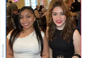 Two women in formal dresses smile for a photo at a gala, one in white, the other in black, holding drinks amid a lively room with branding above.