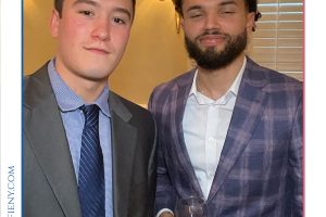 Two men in suits pose for a photo at a formal event, one holding a glass, with a 'Top Lawyers of Long Island' banner above them.