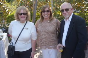Three adults posing outdoors at a social event; two women wearing sunglasses and name badges, man in a dark blazer holding a drink.