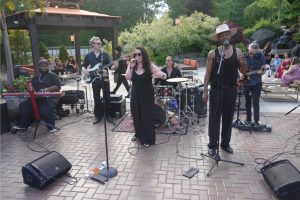 A live band performing outdoors on a brick-paved patio, with a keyboardist, guitarist, drummer, and two vocalists singing into microphones.