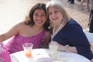 Two women smiling at a sunny outdoor party, one in a pink sequined dress and the other in navy, with drinks on the table in front of them.