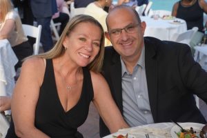 Smiling couple at an outdoor banquet, woman in a black dress with a heart necklace and man in a blazer sit together at a table with plates of food behind them.