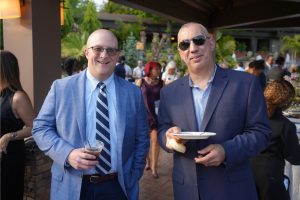 Two men in blue suits pose at an outdoor reception; one holds a cup, the other a plate with bread, both smiling.