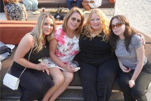 Four women sit closely on an outdoor lounge by the beach, smiling at the camera; two hold drinks and plates are visible nearby.