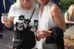 Two smiling women stand together at an outdoor garden party, each holding a cup of beverage.