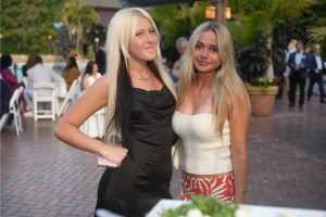 Two young women with long blonde hair posing arm-in-arm at an outdoor event, one in a black dress and the other in a white strapless top and red patterned skirt.