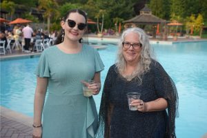 Two smiling women in pastel dresses pose by a pool at an outdoor party, each holding a drink.