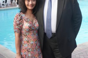Smiling couple posing by a pool at an outdoor event; woman in a pink floral dress with a beige handbag, man in a dark suit and blue tie beside her.