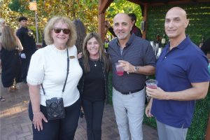 Four adults posing at an outdoor gathering, holding pink drinks at a brick patio bar area.