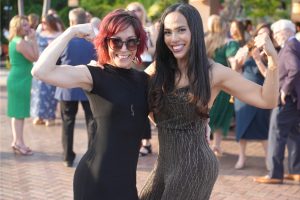 Two women in black dresses flexing their arms and smiling for a photo at an outdoor party, with guests in the background.