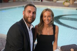 Smiling couple sits poolside, dressed for an evening event, with a man in a suit and a woman in a black dress by the water.