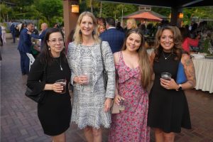 Four women in cocktail attire smiling at the camera at an outdoor social event, each holding a drink.