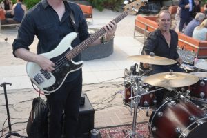 A guitarist with a white bass guitar playing outdoors while a drummer sits at a red drum kit nearby in a garden patio setting with orange couches and lush greenery behind him.