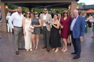 Group of eight adults in semi-formal attire posing together outdoors at a social event, holding drinks.