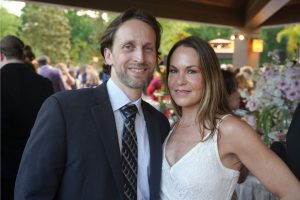 A smiling couple posing closely at a formal event, the man in a dark suit and striped tie, the woman in a white lace dress with a pearl necklace, background guests and floral arrangements.