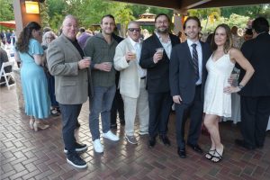 Group of seven adults at an outdoor party, posing with drinks on a brick patio, smiling in smart-casual to formal attire with greenery in the background.