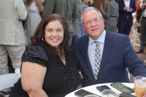 Smiling woman in a black blouse and a man in a blue suit sit at a round table at an outdoor social event, with phones and drinks on the table.