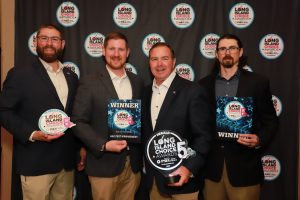 Four men in blazers pose on a step-and-repeat backdrop holding Long Island Choice Awards plaques, smiling after a win vote for 5th year event.